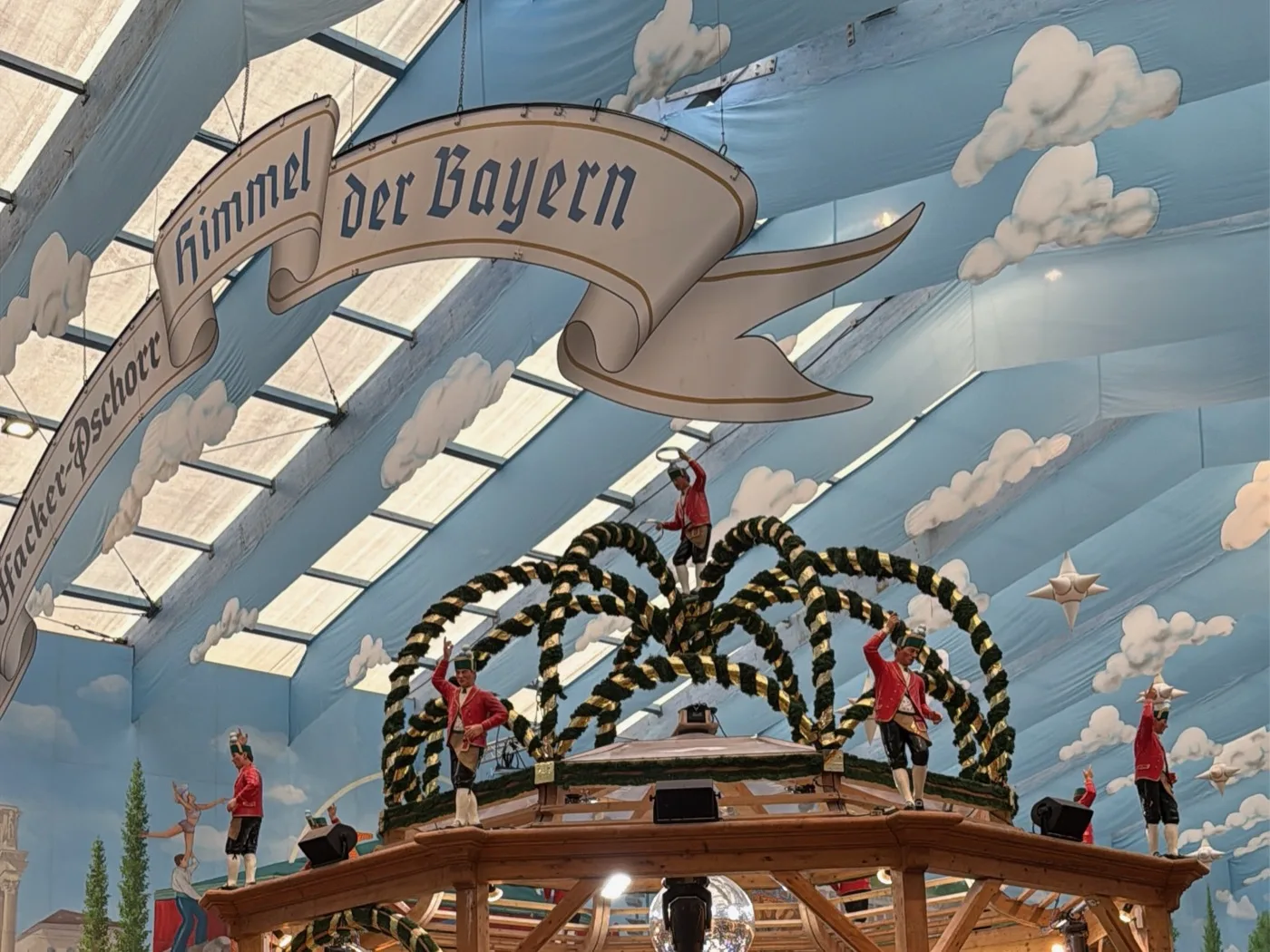 Interior do Hacker-Festzelt: o teto pintado de céu azul com a faixa "Himmel der Bayern" e o palco giratório com a coroa dos Schäffler. Foto minha, setembro de 2026.